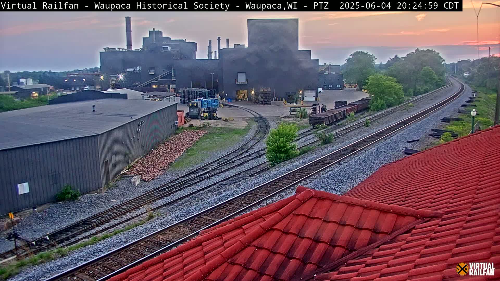 A large industrial building is visible near train tracks at dusk, with a red tile roof in the foreground.