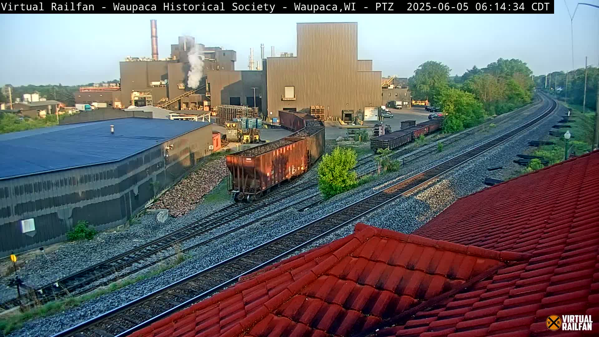 A railway with several train cars sits near an industrial complex on a partly sunny day.