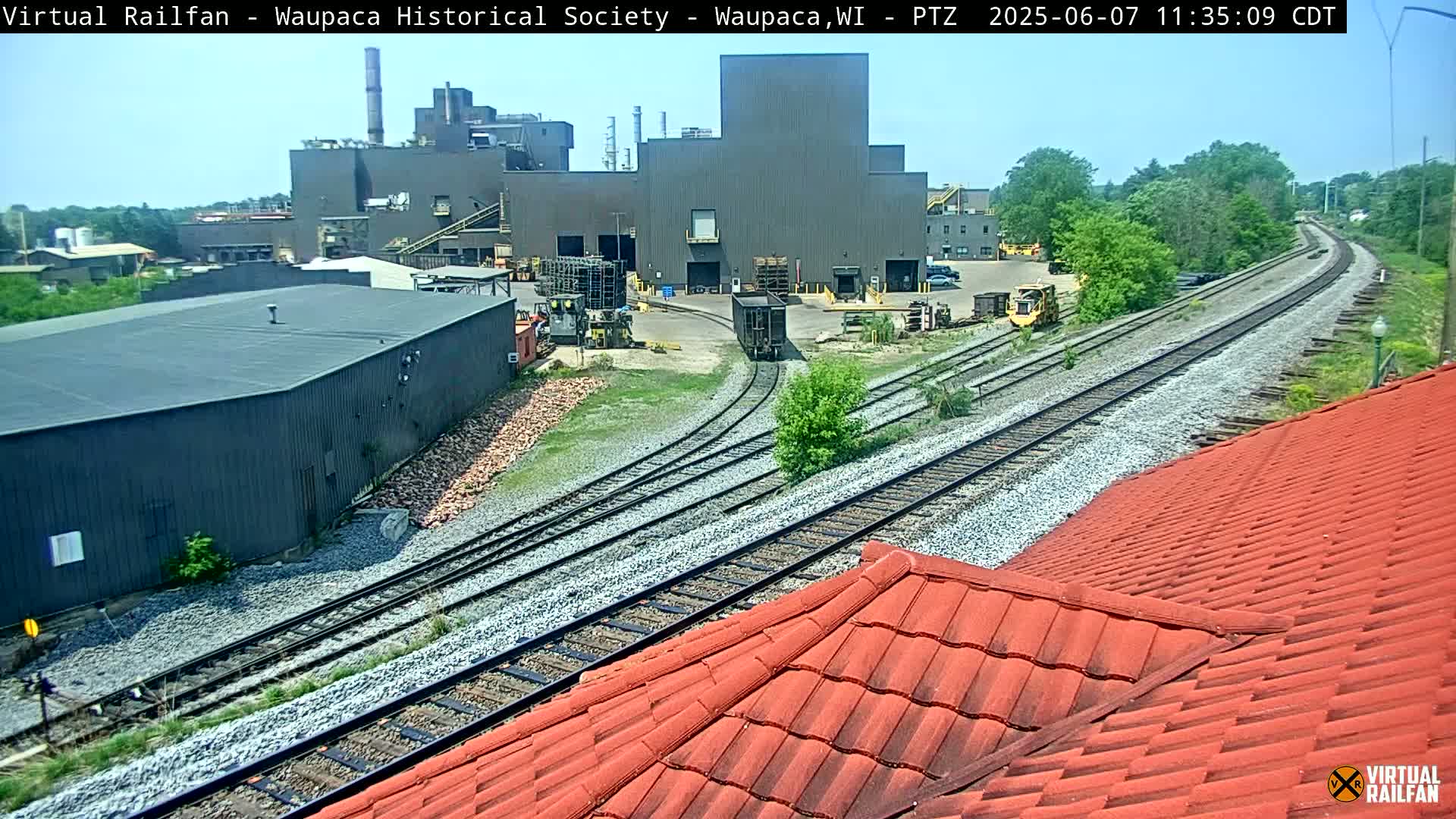 A section of railway tracks curves past an industrial complex on a sunny day, partially visible from a red tiled roof.
