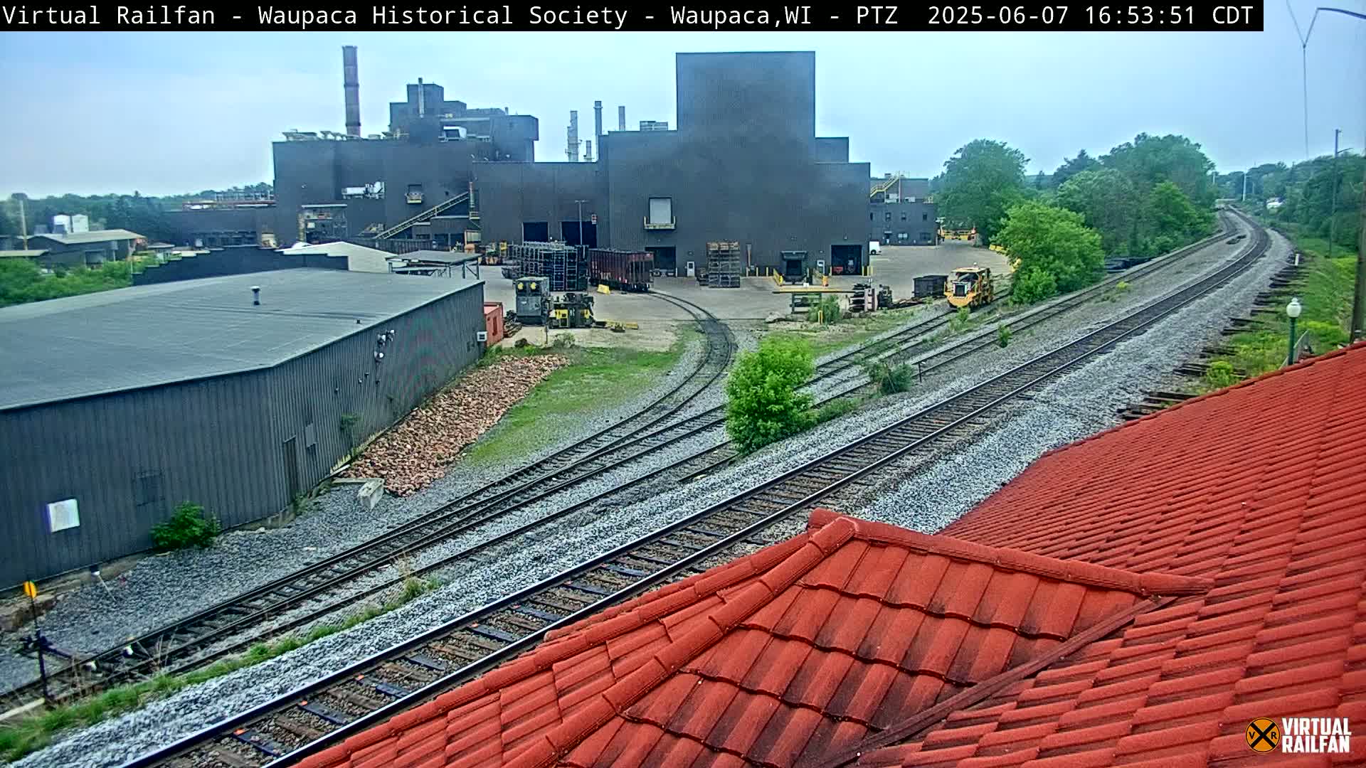 A view of railroad tracks curving past industrial buildings and a red tile roof under an overcast sky.