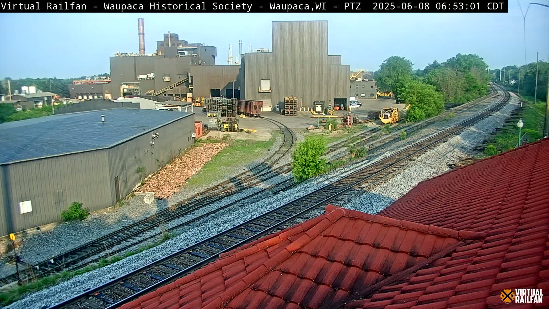 Under a clear sky, railroad tracks curve past industrial buildings and a red-tiled roof.