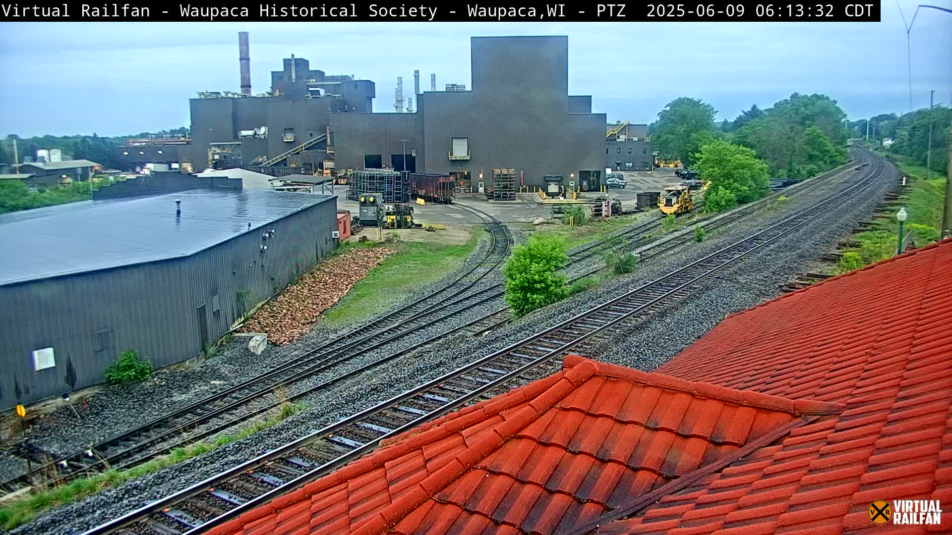 A large industrial building is situated near a railway, with overcast skies and a red-tiled roof partially visible in the foreground.