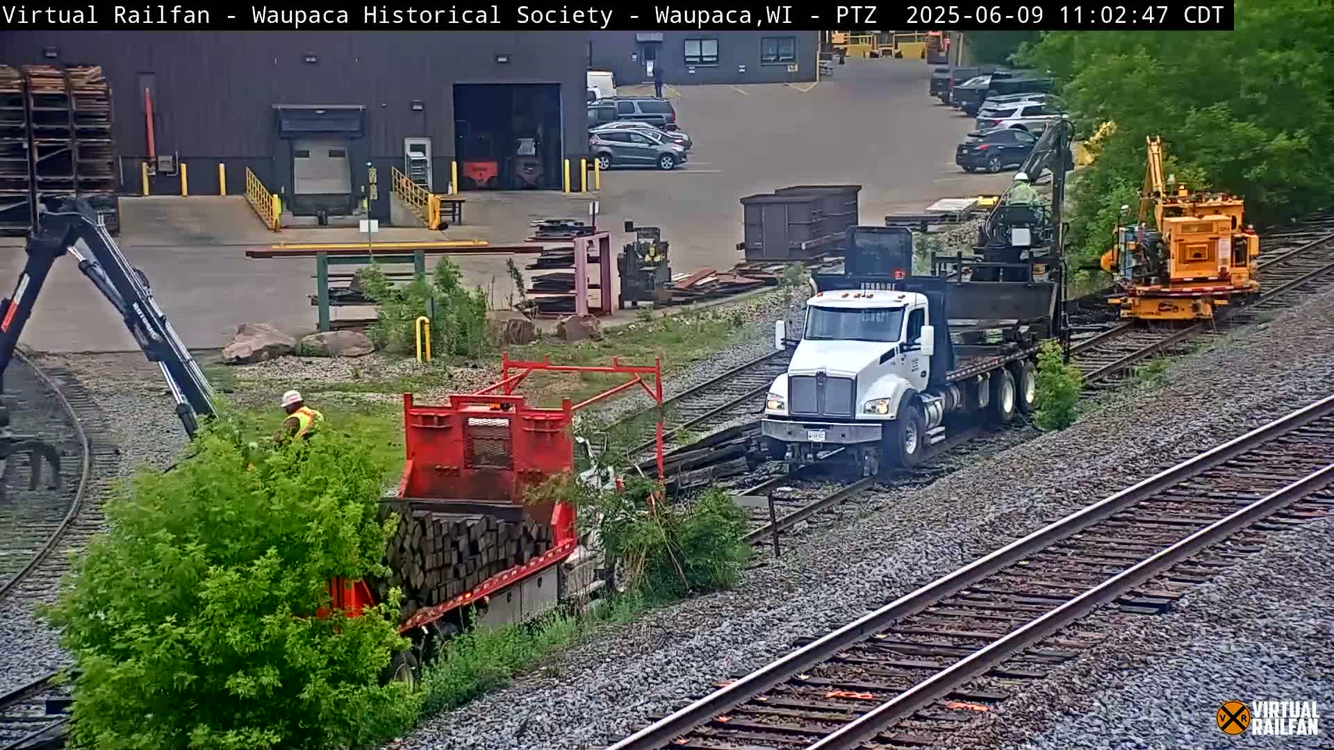 A white semi-truck and a red flatbed truck are parked on railroad tracks near a factory; a worker is loading a flatbed truck with rectangular objects, next to some bushes, in an overcast outdoor setting.