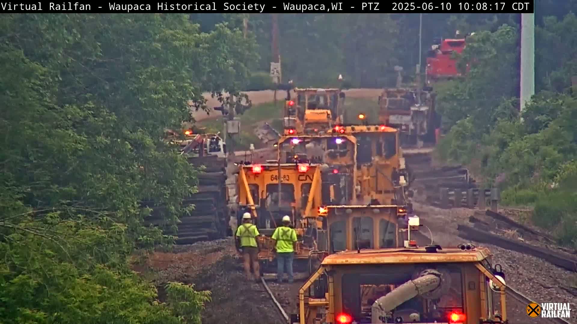 A line of yellow railway maintenance vehicles is on the tracks, with two workers in yellow vests visible near the center, under an overcast sky.