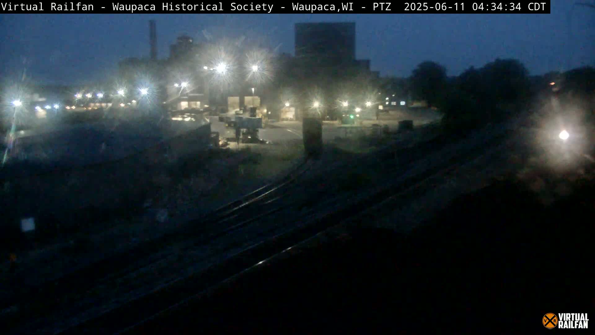 A nighttime view of a dimly lit industrial area with railroad tracks in the foreground under a dark sky.