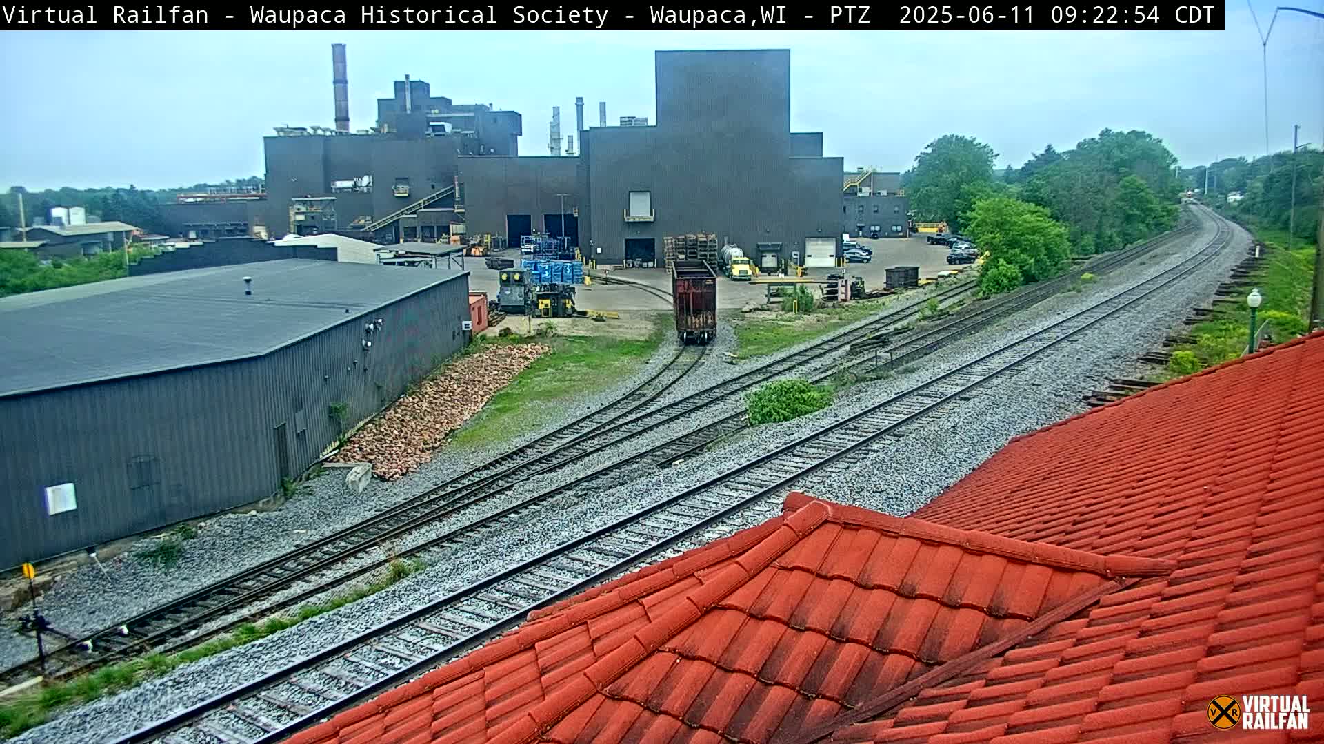 A rail yard with a large industrial building and a red tiled roof in the foreground under an overcast sky.