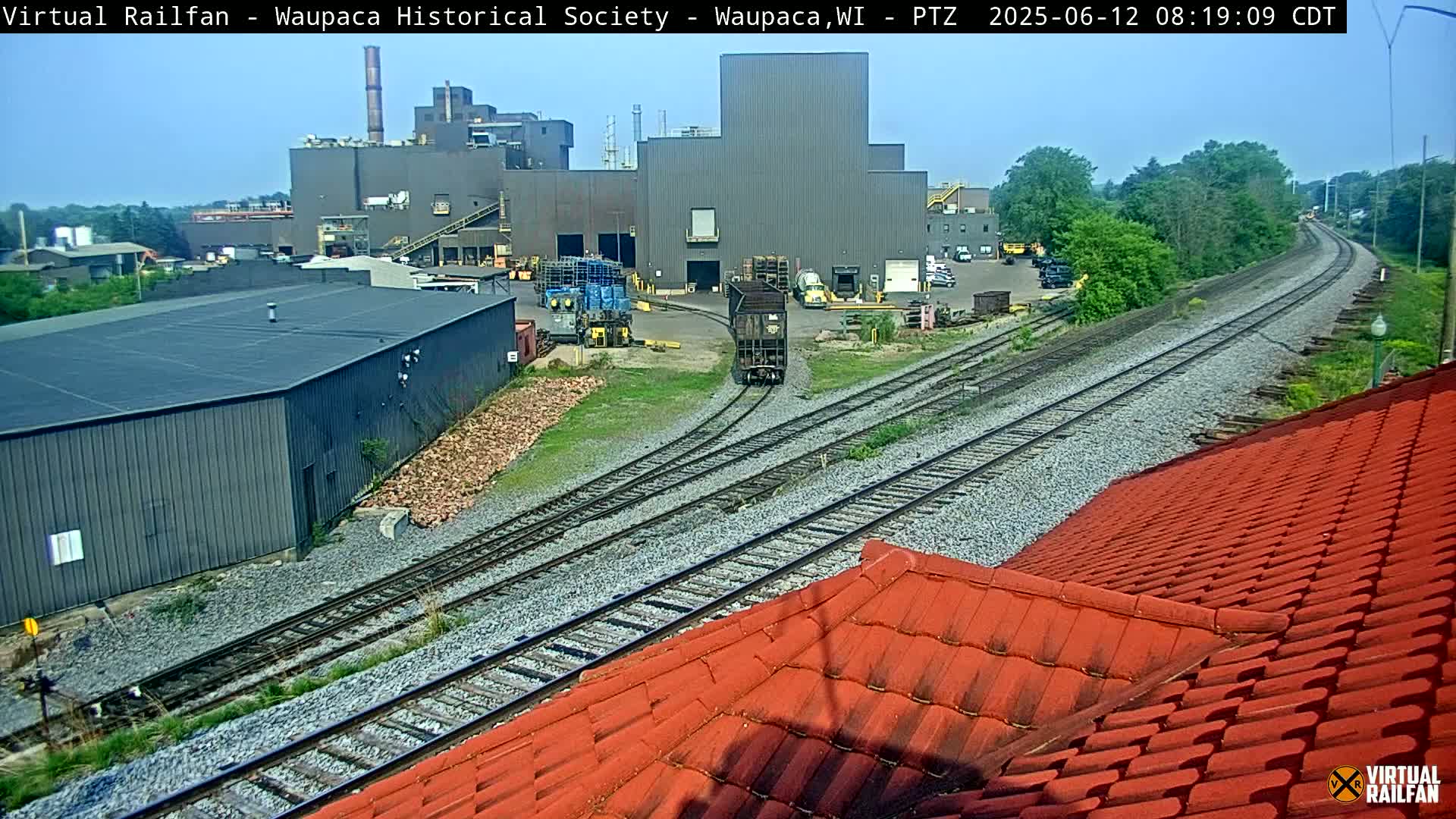 A train car passes an industrial complex on a sunny day near a section of railway tracks.
