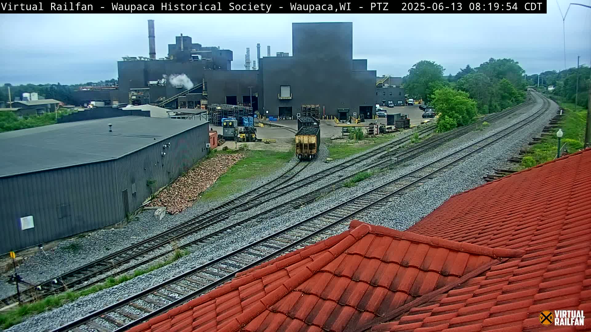 A railcar moves along train tracks near an industrial building complex on an overcast day.
