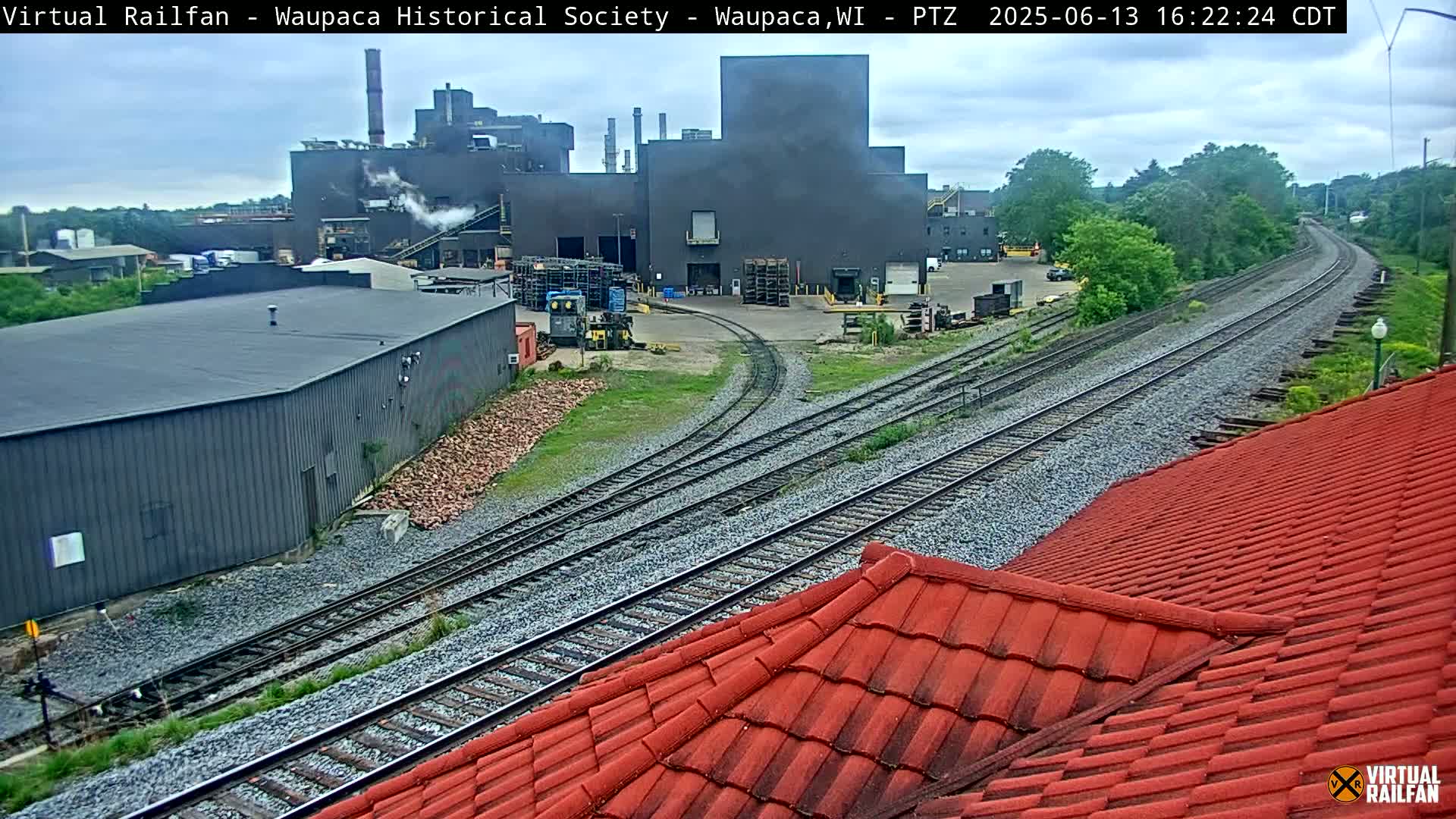 A large industrial building with smokestacks is visible in the background, alongside a railway system with converging tracks under an overcast sky.