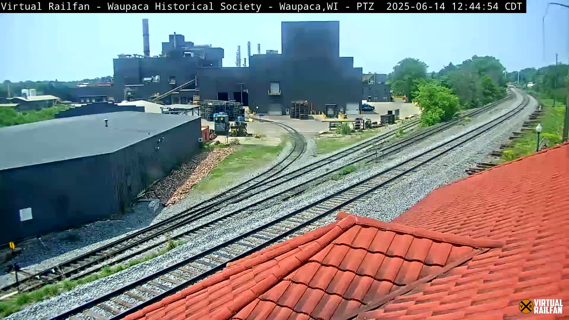 A large industrial building complex is visible next to railroad tracks on a sunny day.