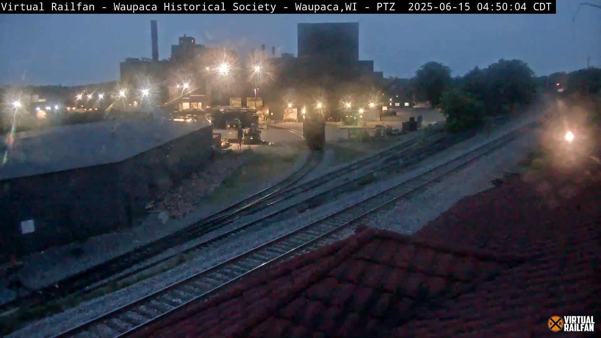 A nighttime view of a large industrial building and railroad tracks, under a dark, possibly twilight sky.