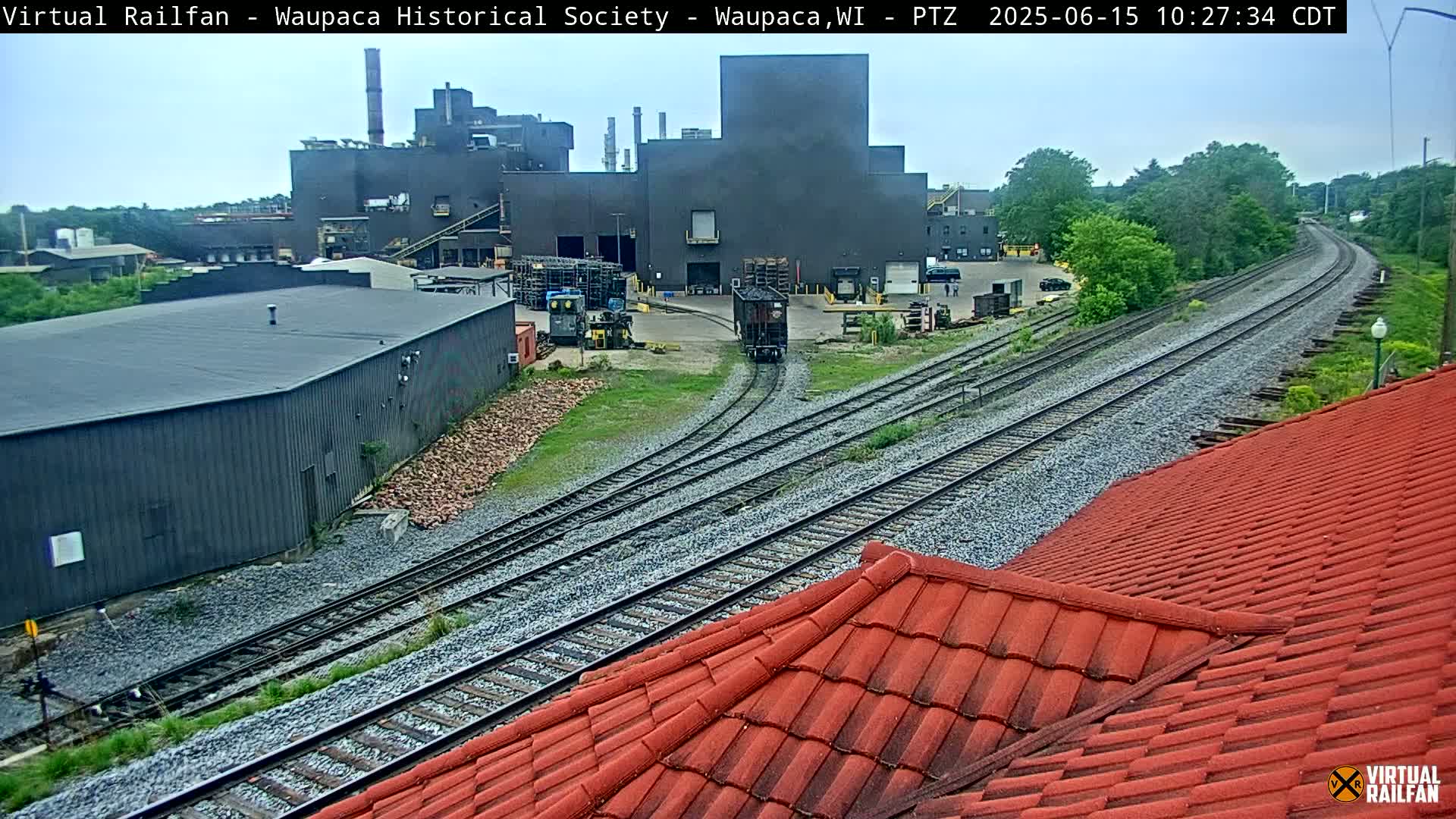 A train moves along railway tracks that pass near an industrial building complex on an overcast day.