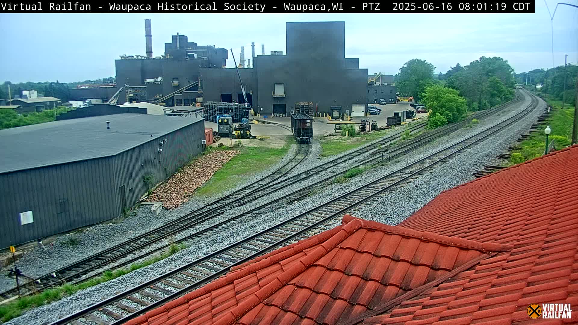 A single rail car travels along train tracks near a large industrial building complex on an overcast day.