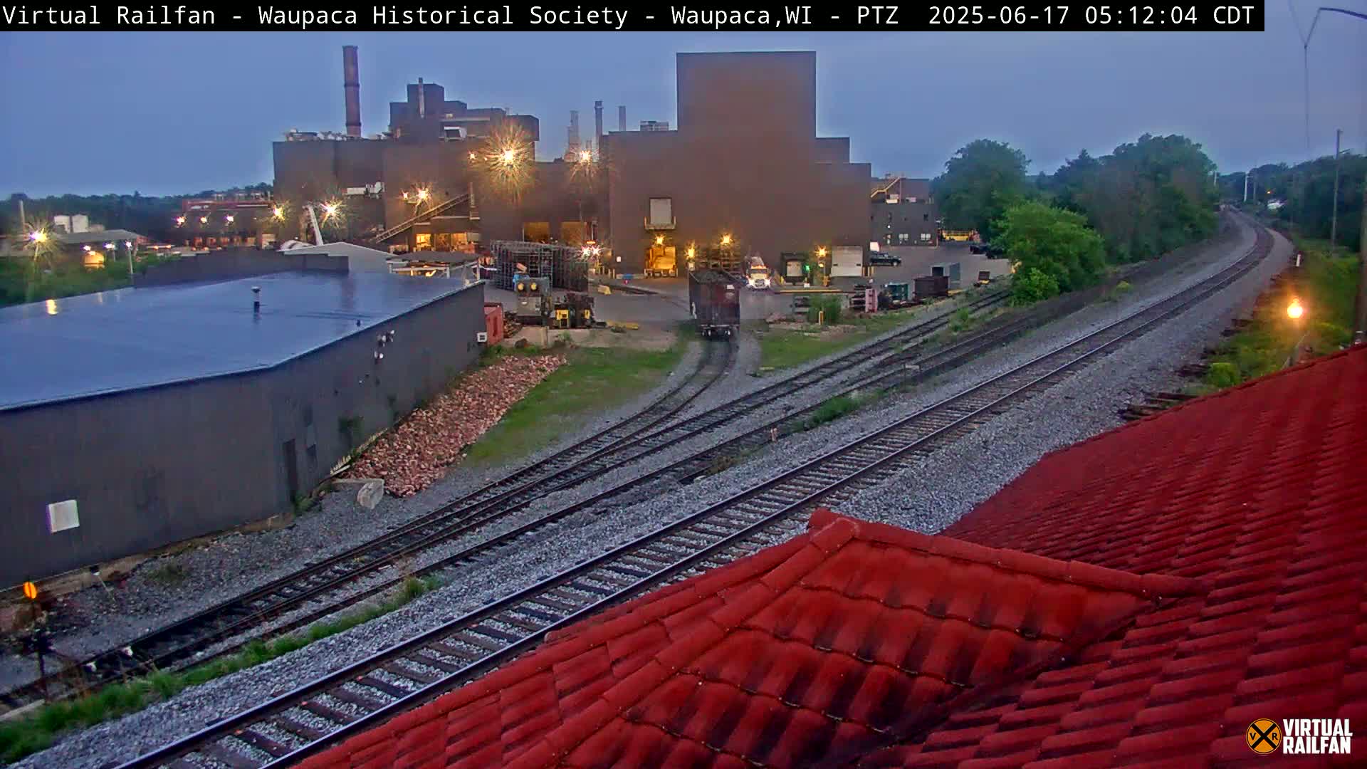 A train car approaches a rail yard near a large industrial building under a twilight sky.