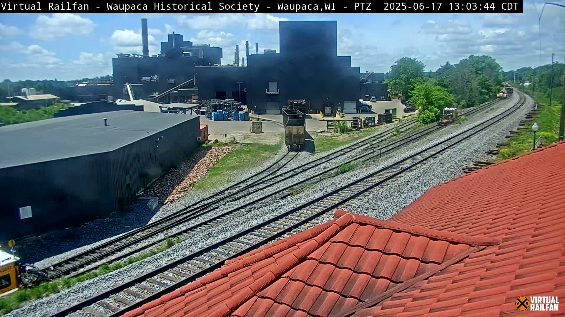 A partly sunny day shows a large industrial building near train tracks with several railcars visible, including one moving on a switch.