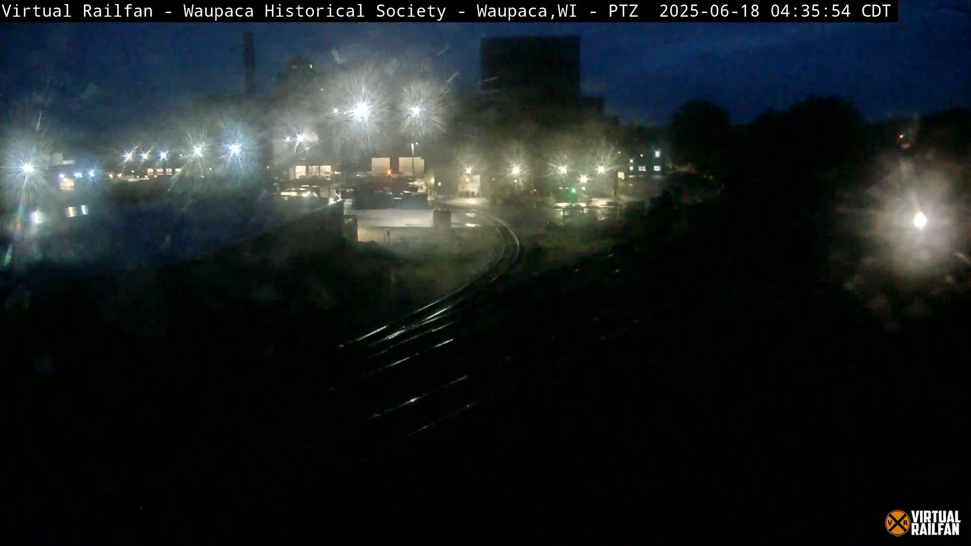 A nighttime, rain-soaked view of railroad tracks curving past illuminated buildings and trees.