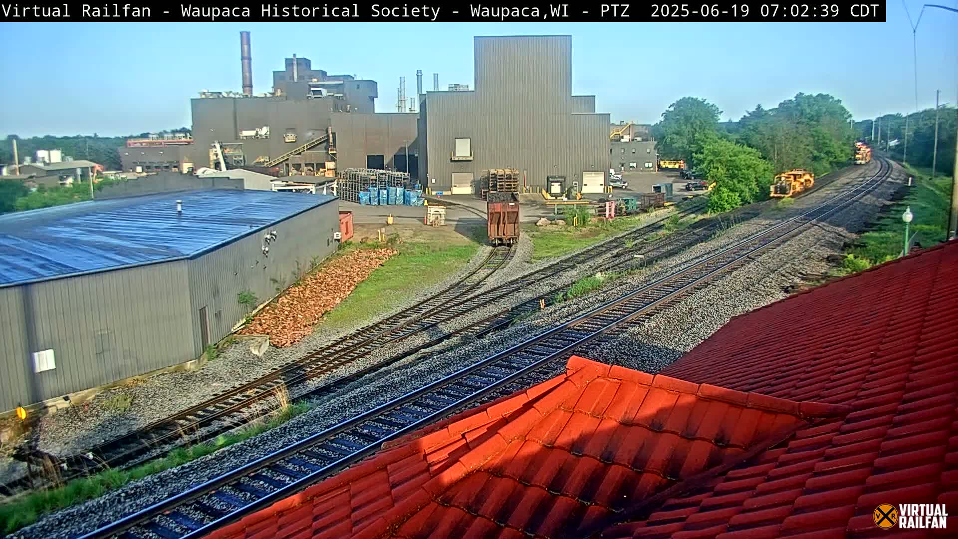 A red-tiled roof is in the foreground, overlooking railroad tracks with a train car and industrial buildings under a sunny sky.