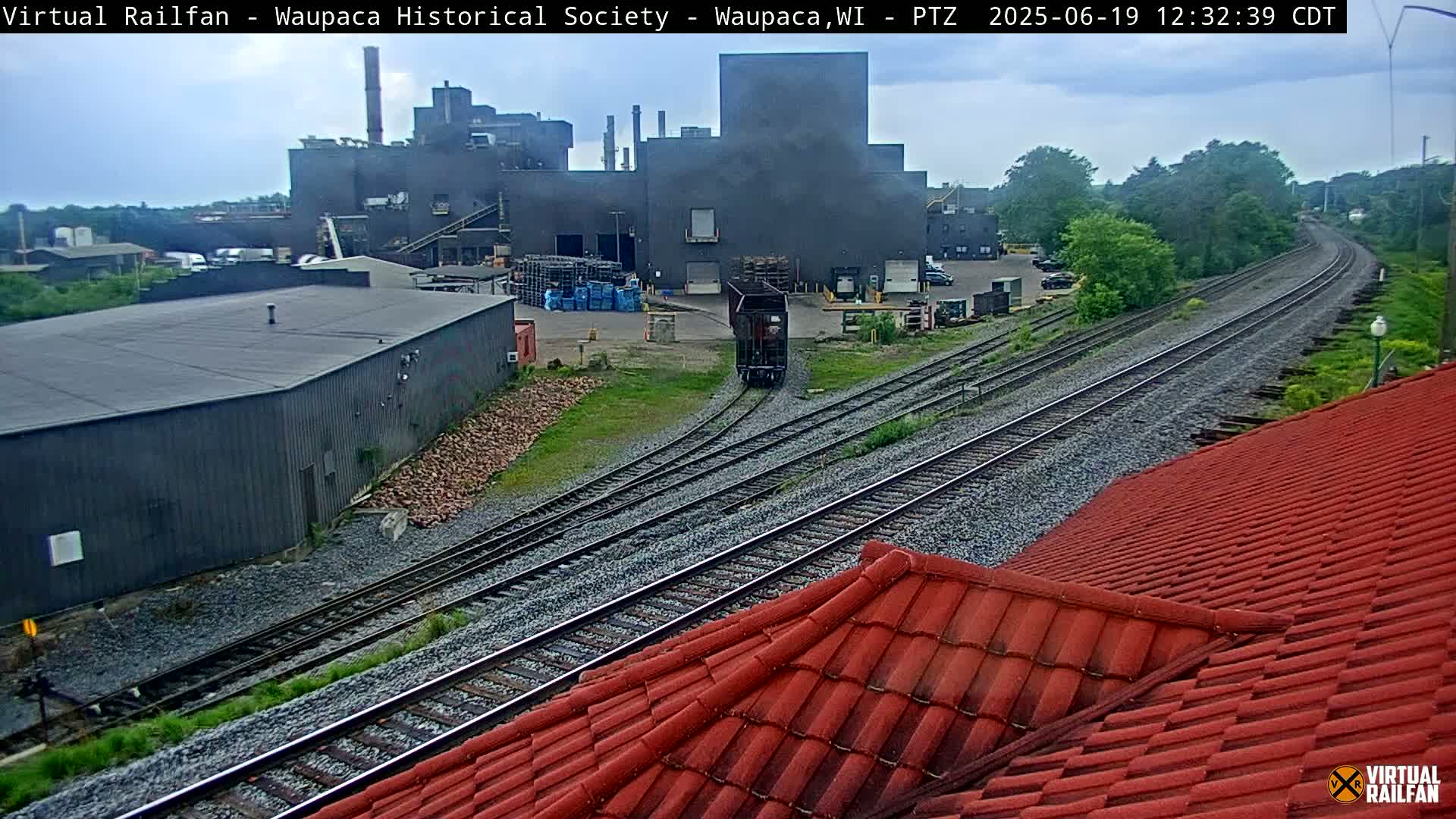 A train travels along railroad tracks toward a large industrial building complex under a partly cloudy sky.