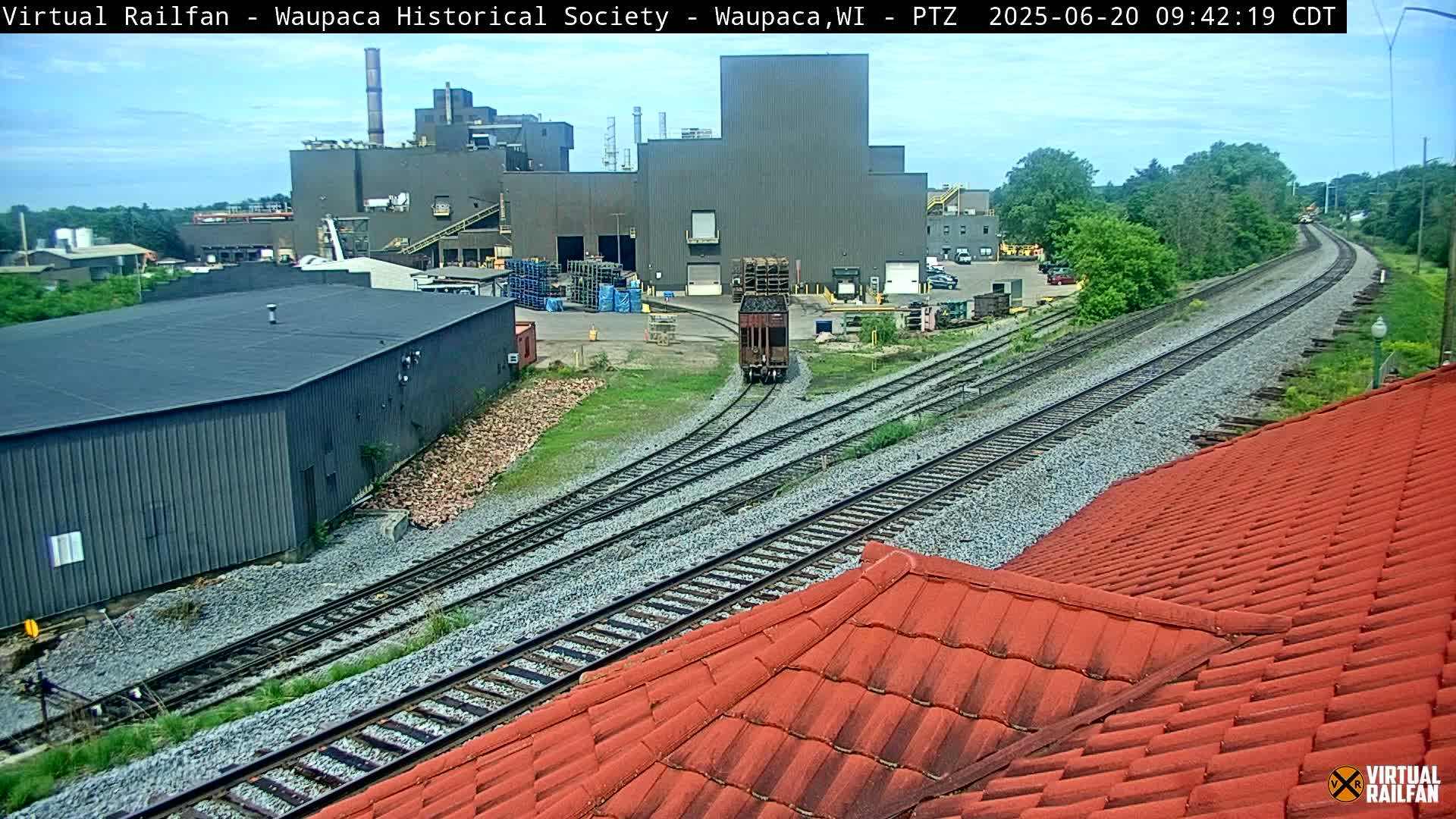 A railcar moves along train tracks near an industrial building on a partly cloudy day.
