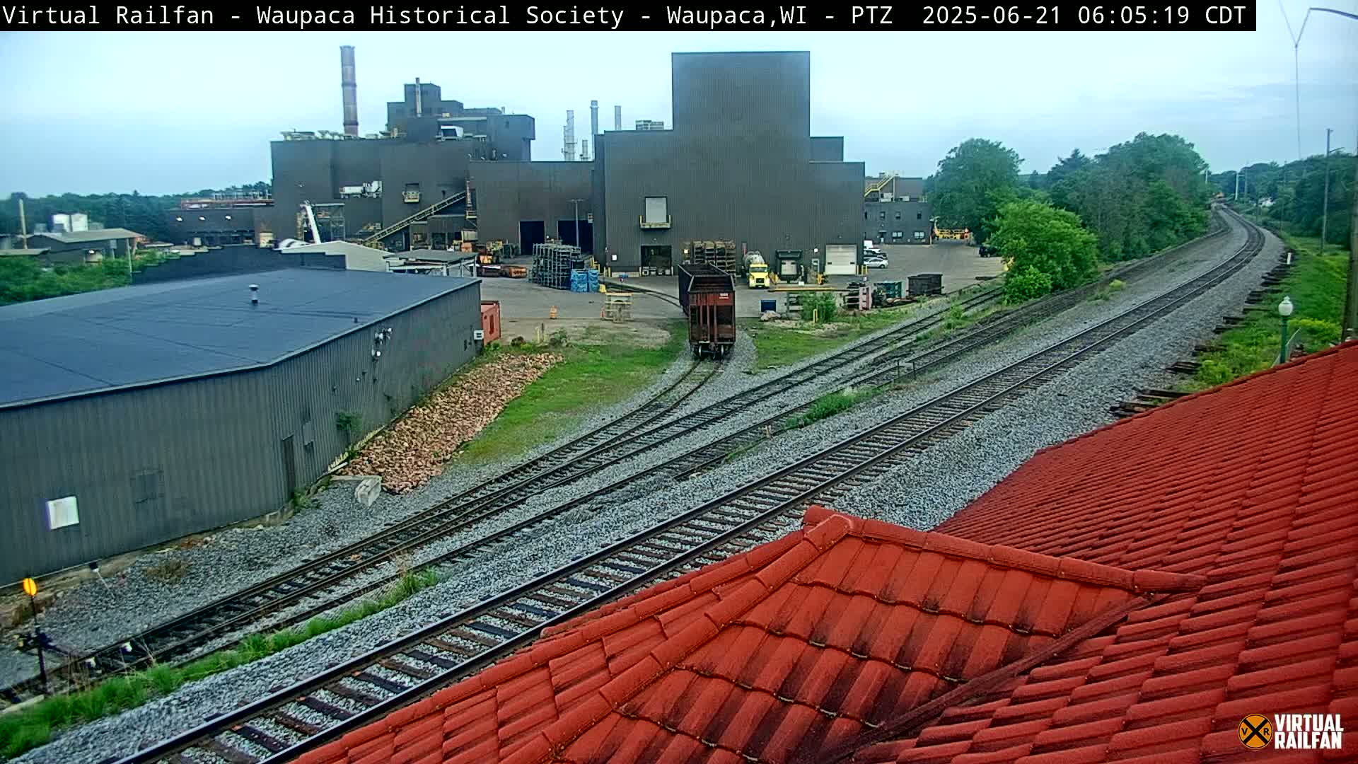 A railroad car travels along a track toward a large industrial building under an overcast sky.