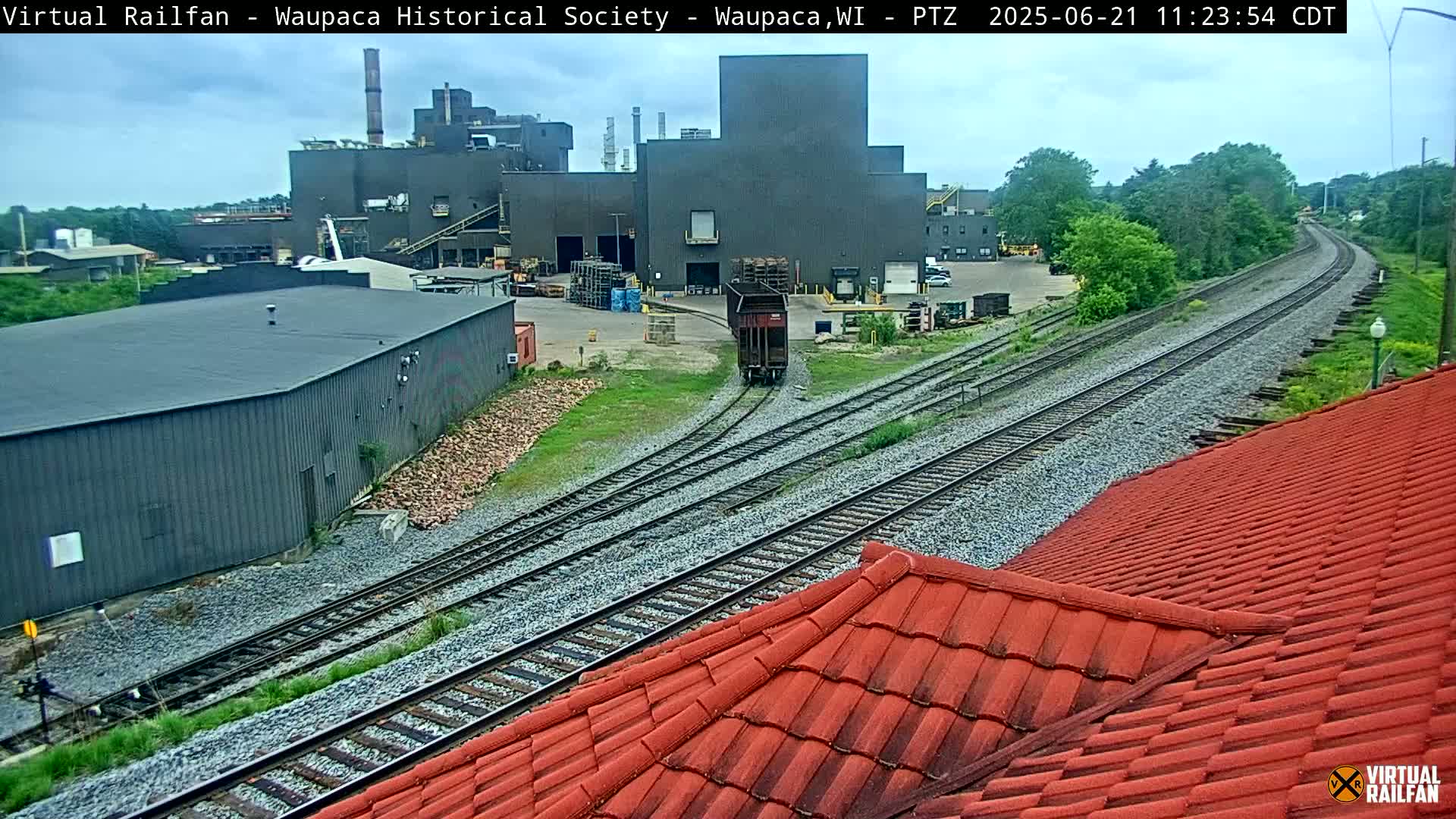 A single freight car travels along railway tracks past an industrial complex on an overcast day.
