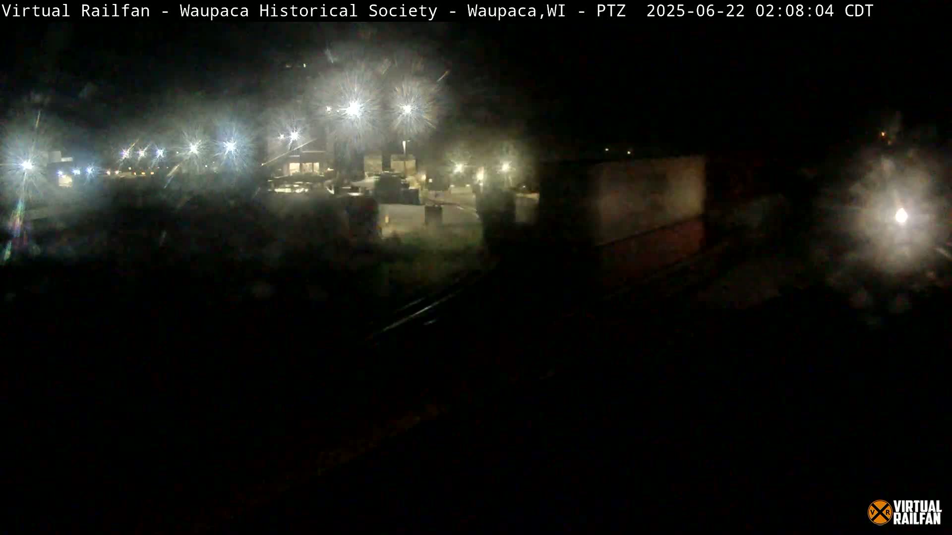 A nighttime, low-light view of a rail yard with several illuminated buildings and tracks visible in the distance.