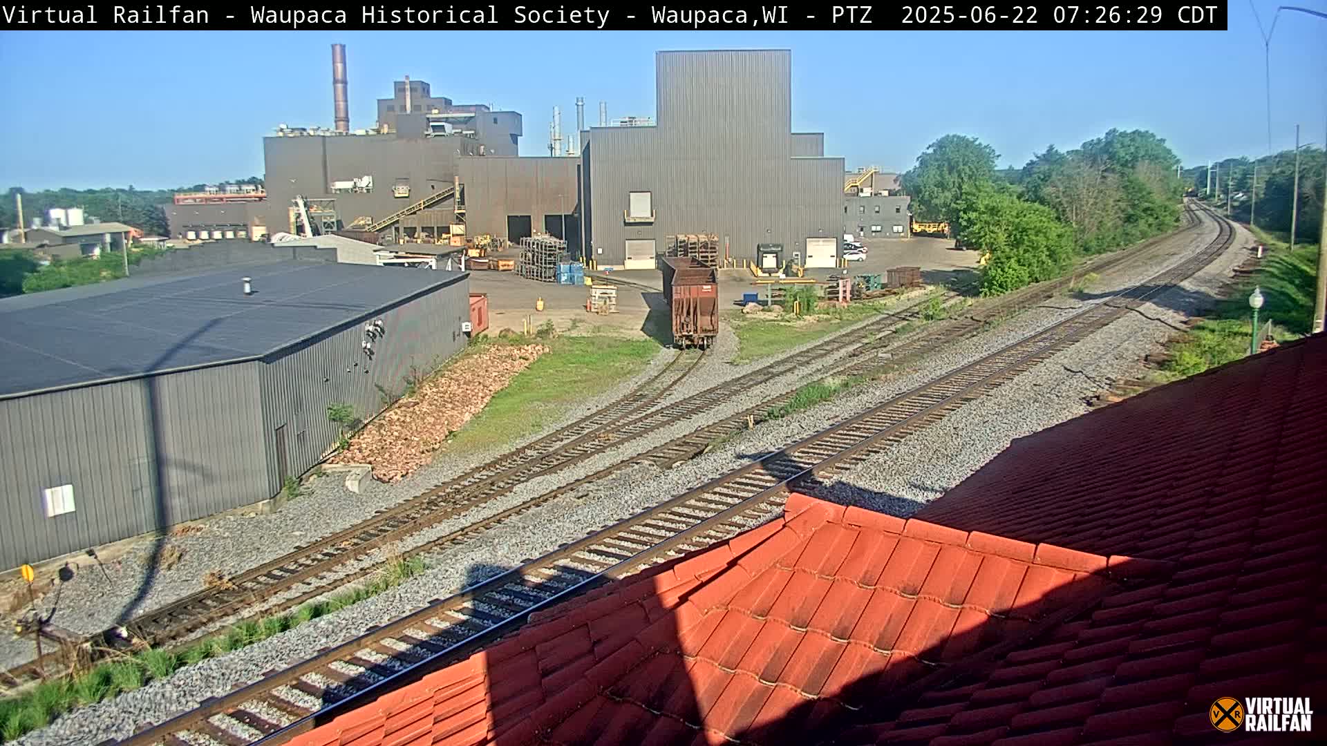A railway line curves past an industrial complex on a sunny day, with a single rail car visible nearby.
