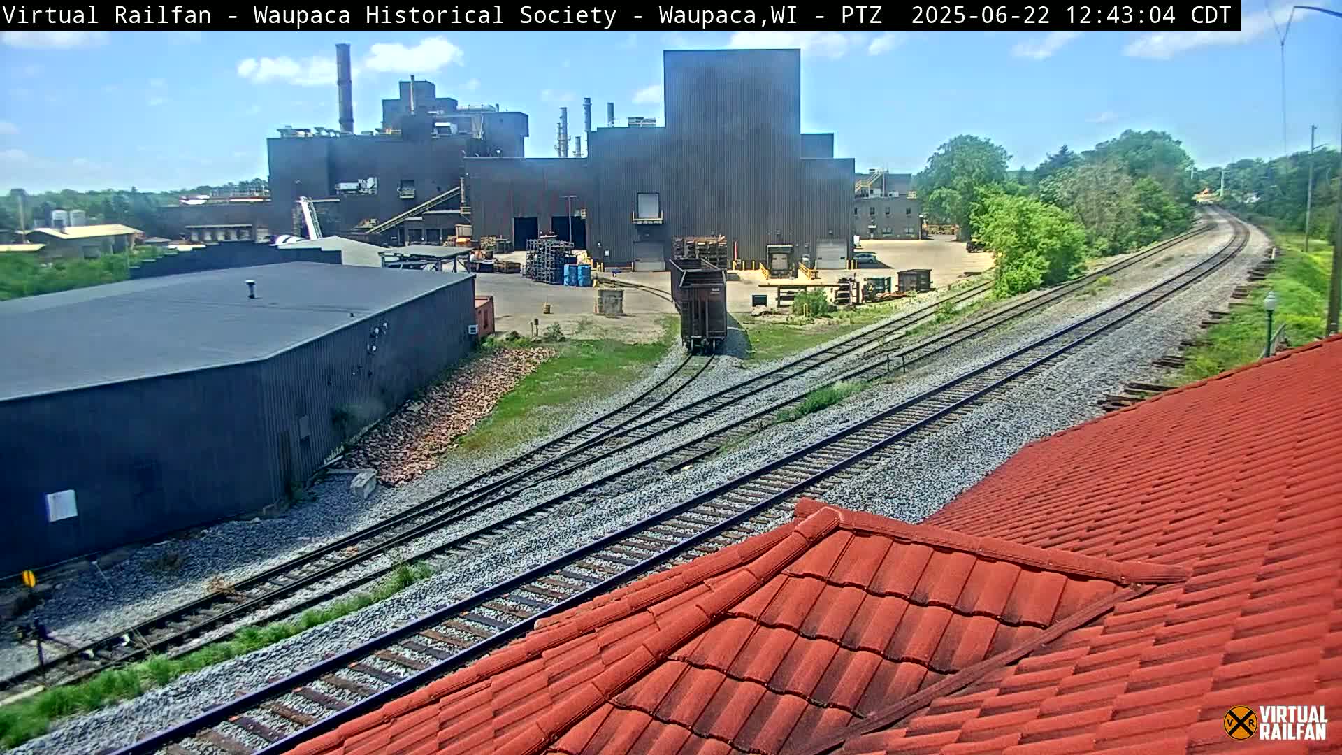 A rail yard with a large industrial building and a red tiled roof in the foreground under a partly sunny sky.