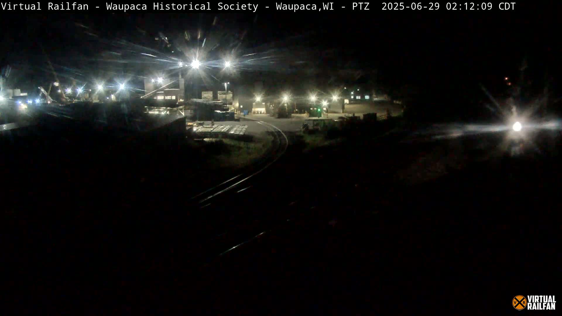 A nighttime view of an industrial area with railroad tracks, illuminated by several bright lights.