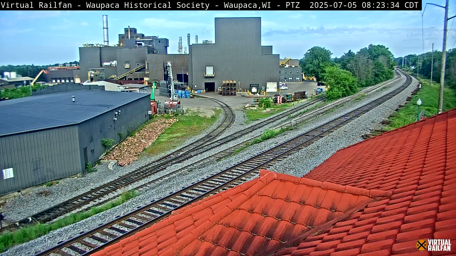 A large industrial building complex is situated beside a railway track on a partly cloudy day.