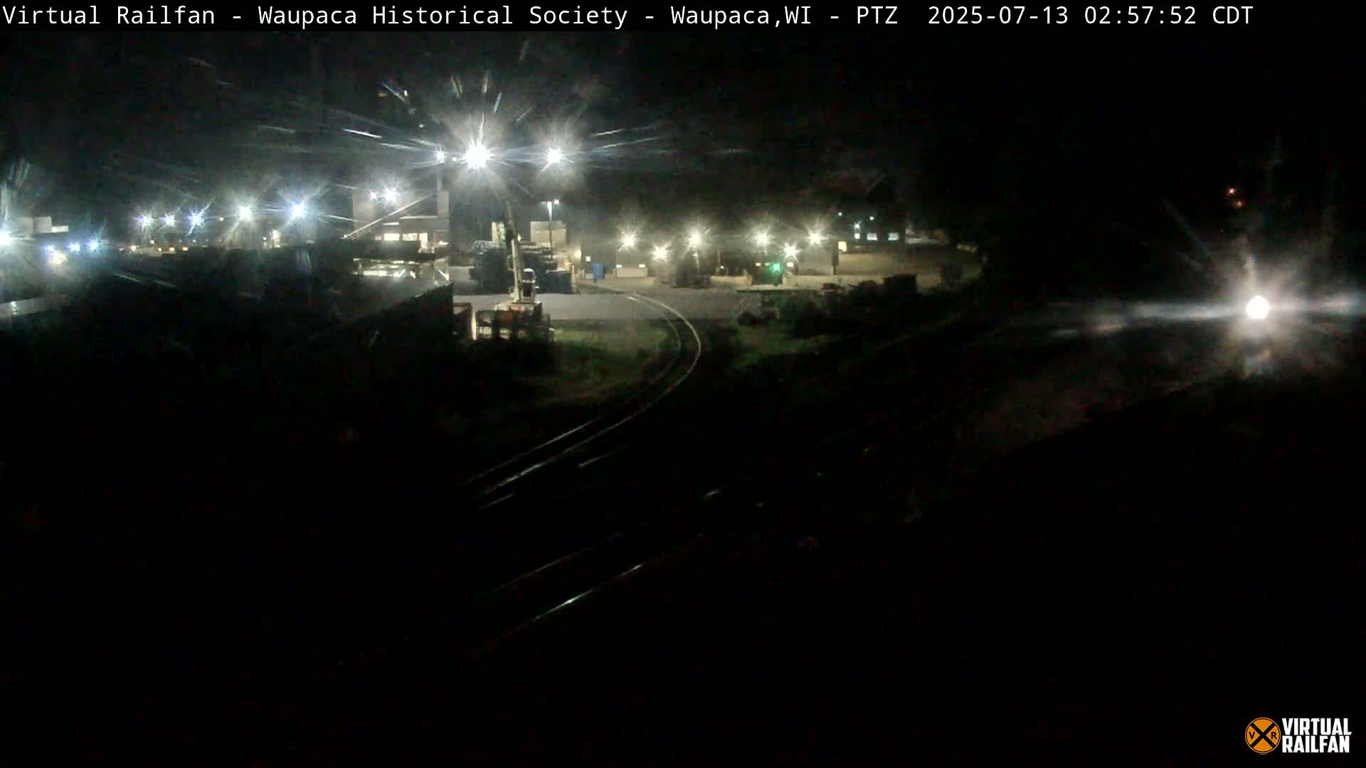 A nighttime view of a dimly lit train yard with railroad tracks and several lights illuminating the area.