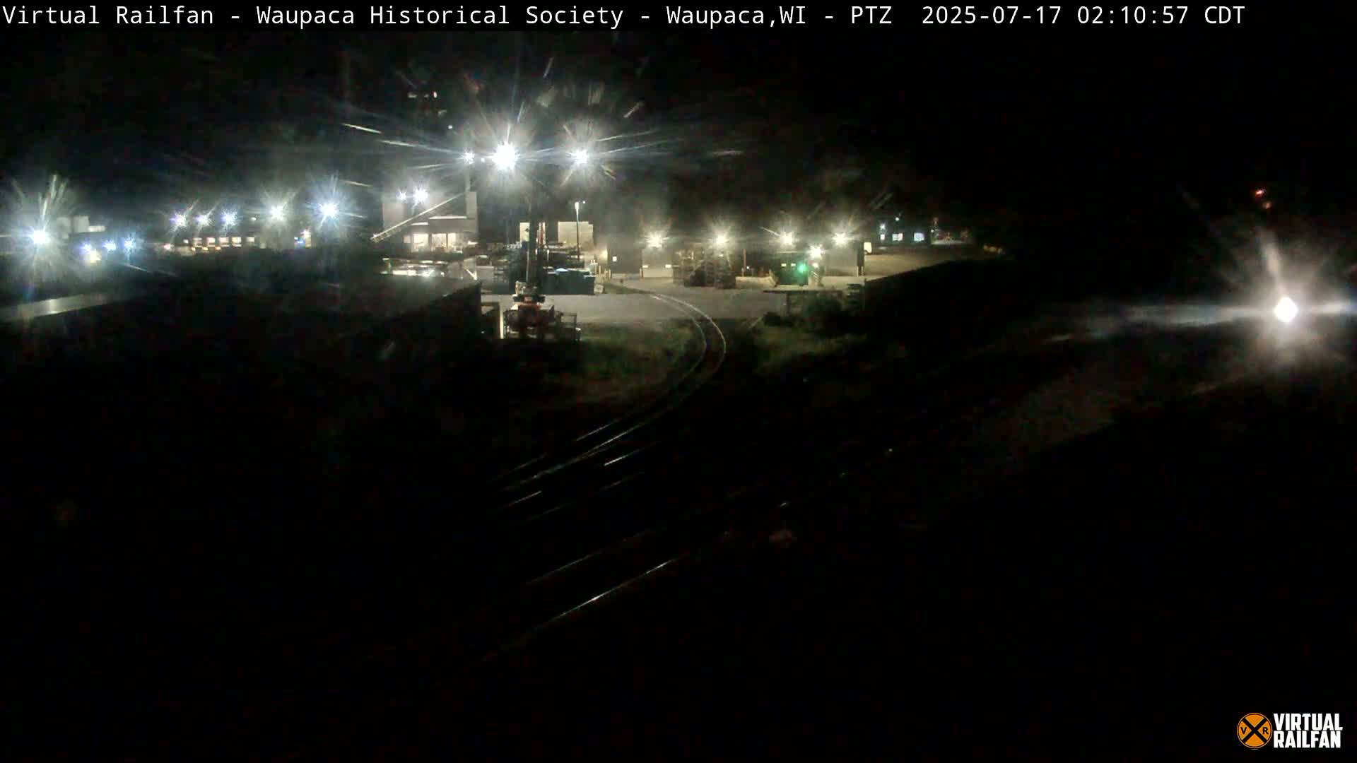 At night, brightly lit industrial buildings and railway tracks are visible under a clear, dark sky.