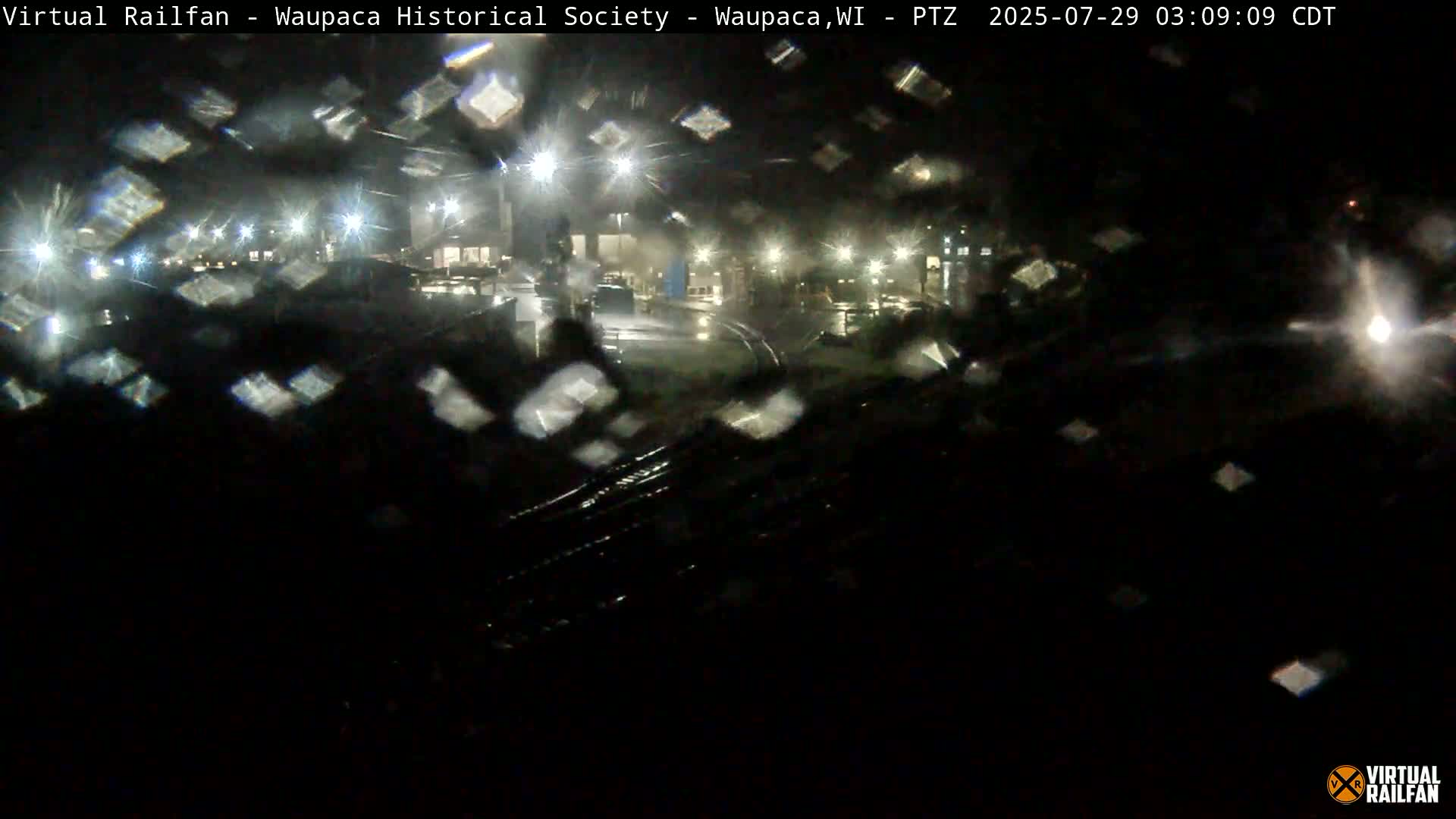 A nighttime view of a wet, dimly lit area, possibly a parking lot or industrial site, seen through a rain-streaked window.