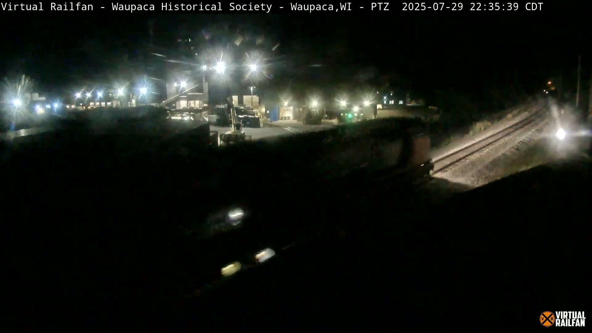 A nighttime view of a wet, dimly lit area, possibly a parking lot or industrial site, seen through a rain-streaked window.