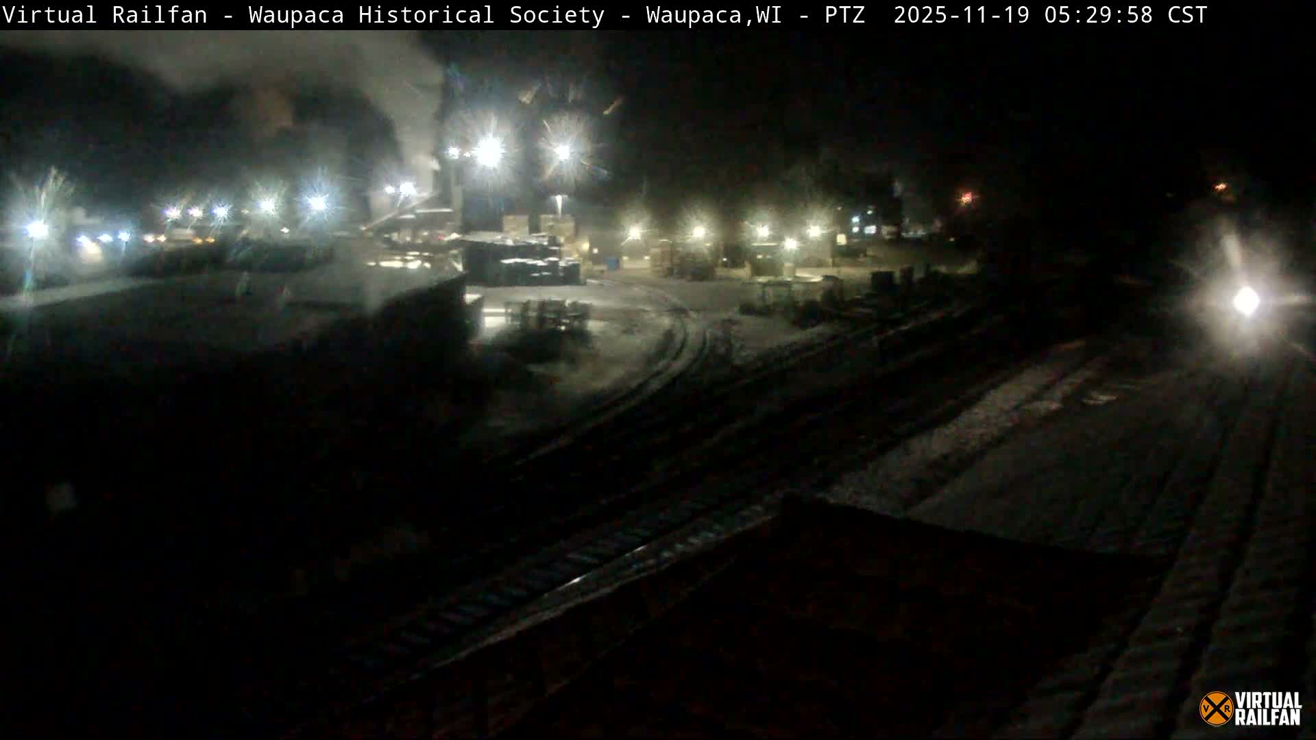 A nighttime view of a wet, dimly lit area, possibly a parking lot or industrial site, seen through a rain-streaked window.