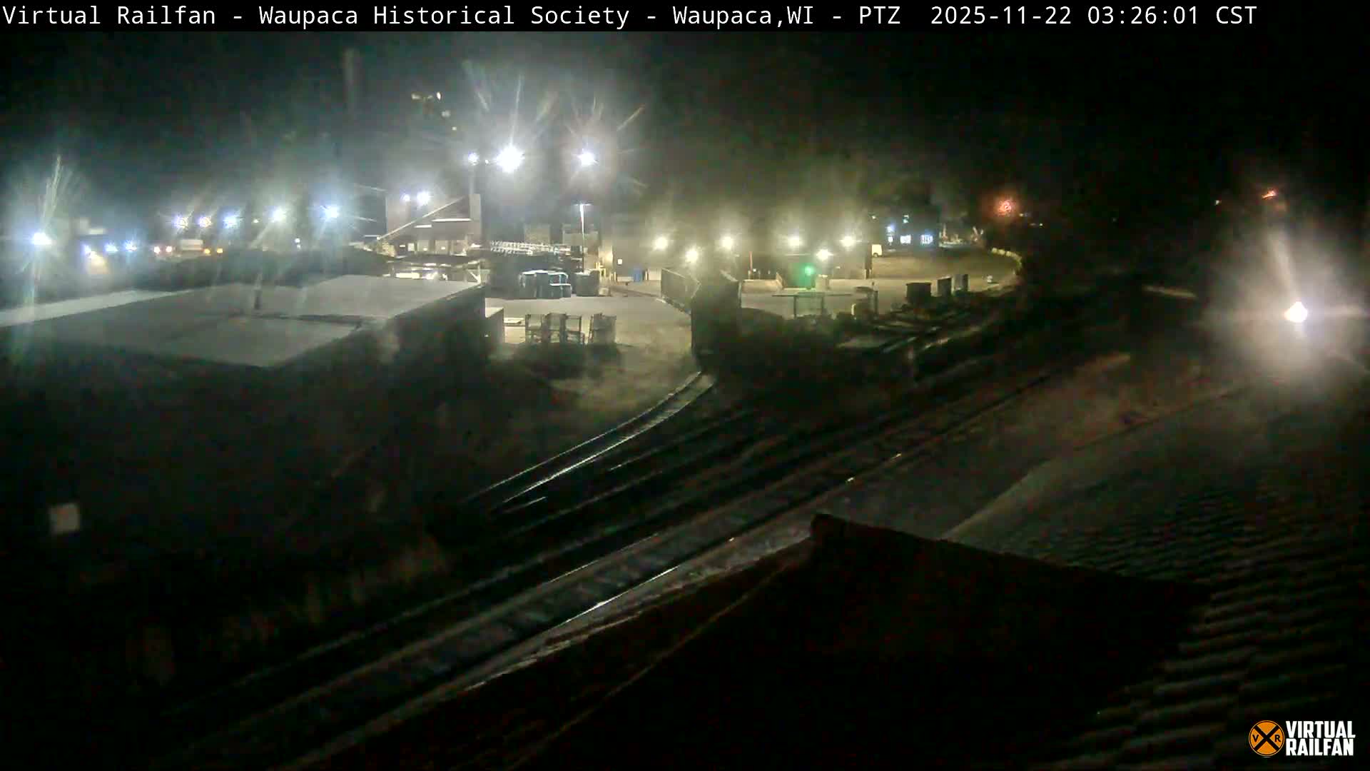Under clear night skies, an illuminated railyard or industrial area is visible, featuring multiple train tracks in the foreground, buildings and stacked materials in the mid-ground, and numerous bright artificial lights, with a prominent headlight on the far right.
