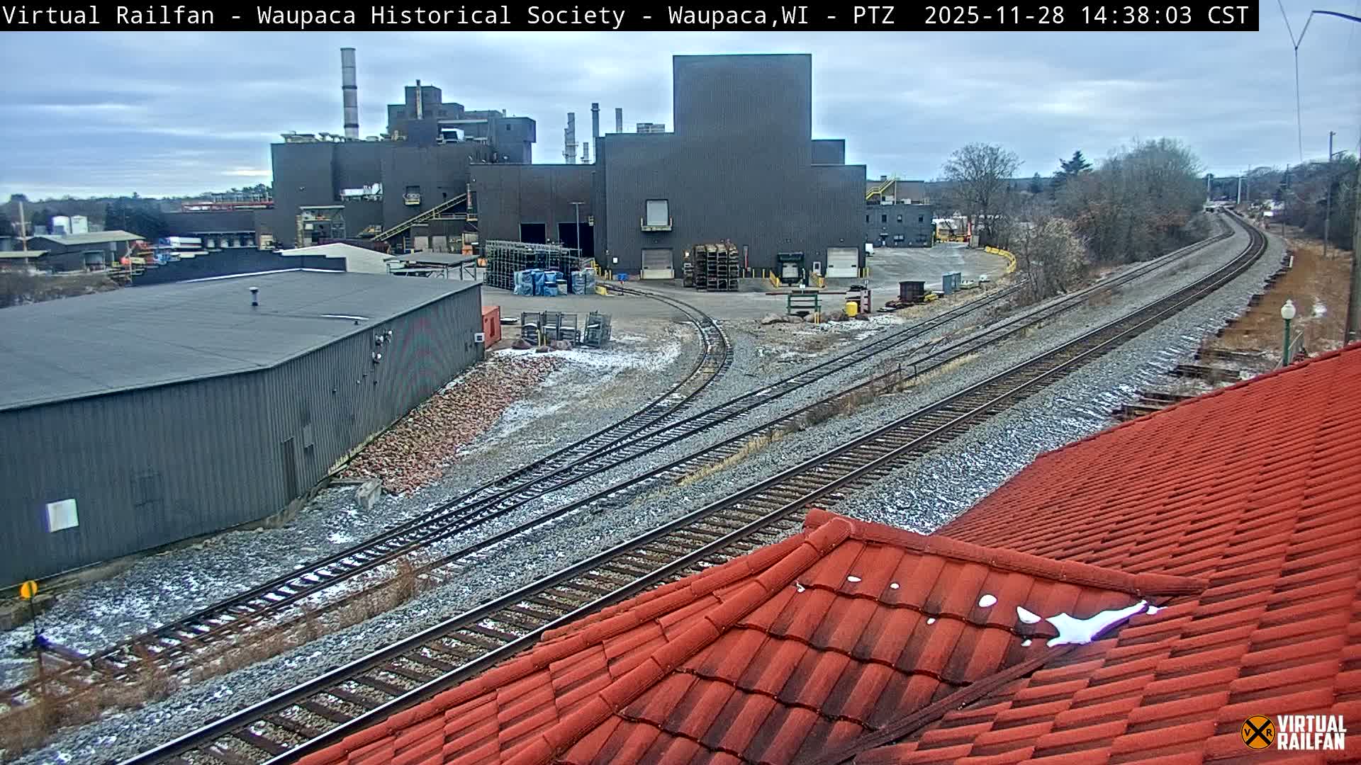 Under an overcast sky, an industrial rail yard features multiple snow-dusted tracks curving past large dark gray buildings, with a red tile roof showing patches of snow dominating the foreground.