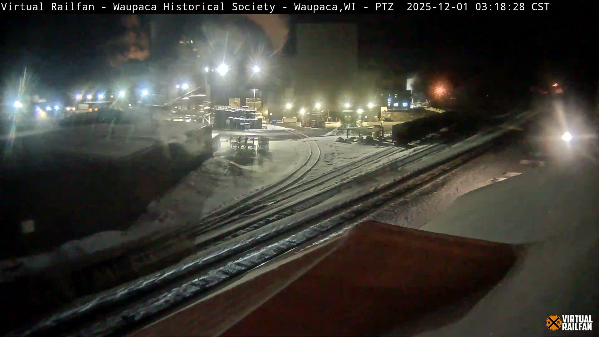 A snow-covered railroad yard is seen at night, with multiple tracks illuminated by streetlights and building lights, and a bright light visible in the distant right.