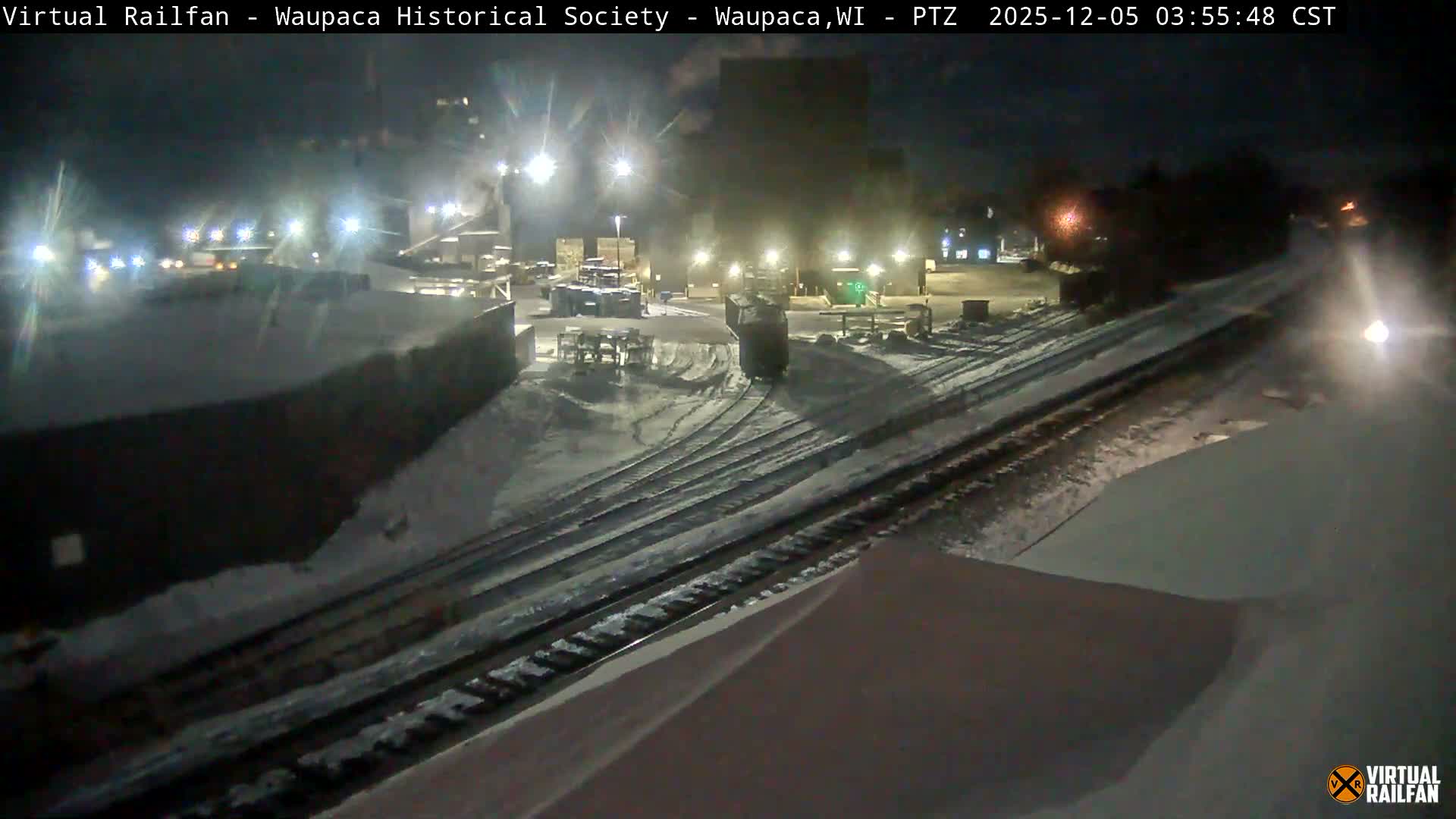 A snowy nighttime scene displays multiple train tracks illuminated by bright lights, with a distinct powerful headlight visible in the distance along the tracks.