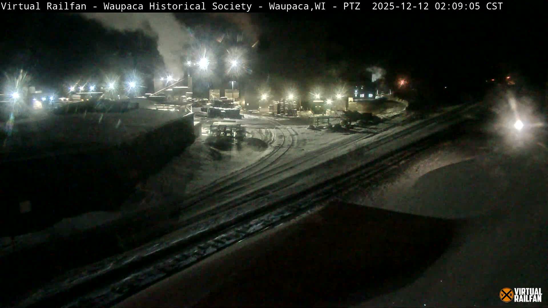 A snow-covered industrial complex or rail yard is illuminated by bright lights at night, with steam rising from structures and a train approaching in the distance.