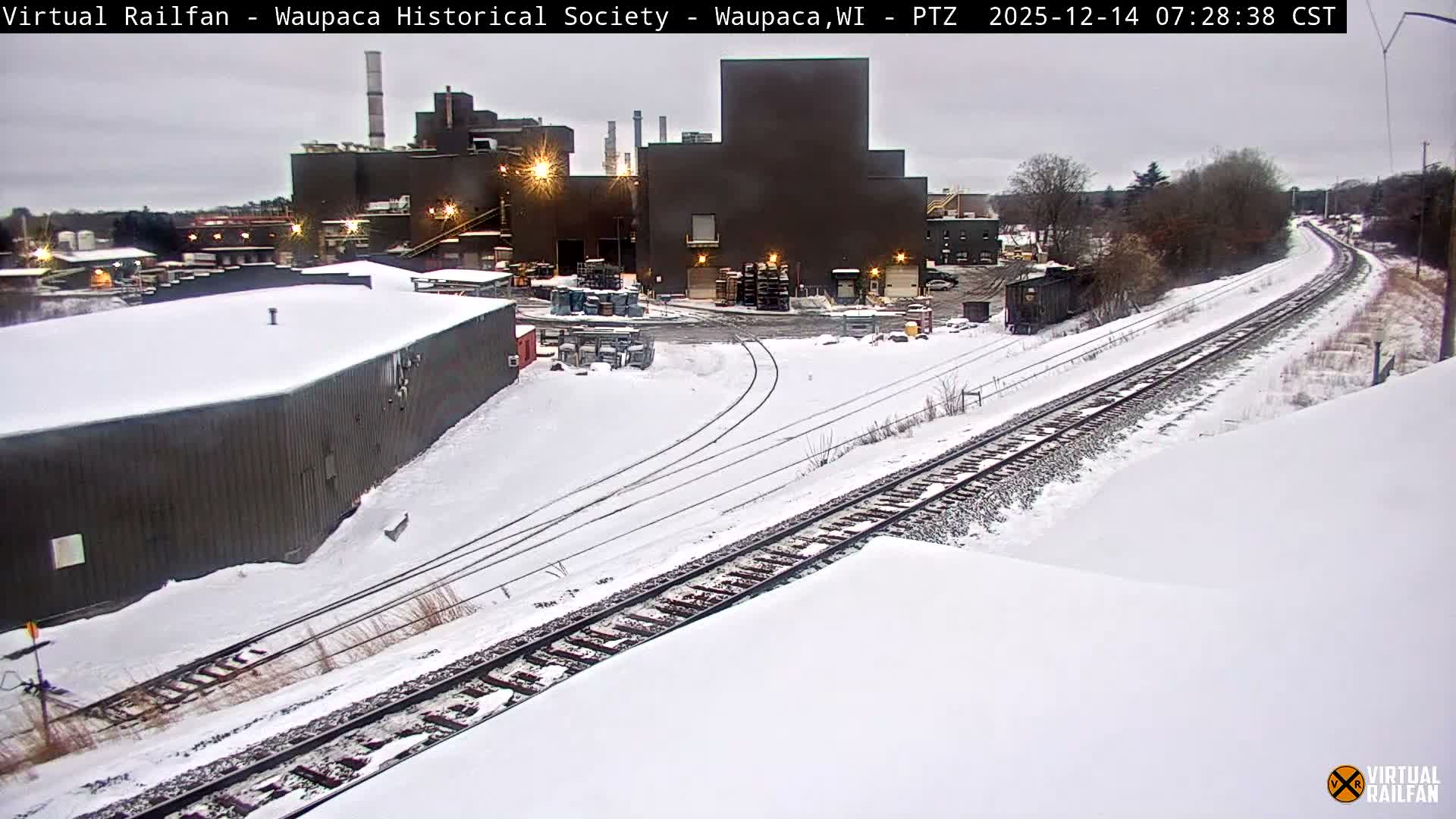 A snow-covered industrial complex or rail yard is illuminated by bright lights at night, with steam rising from structures and a train approaching in the distance.