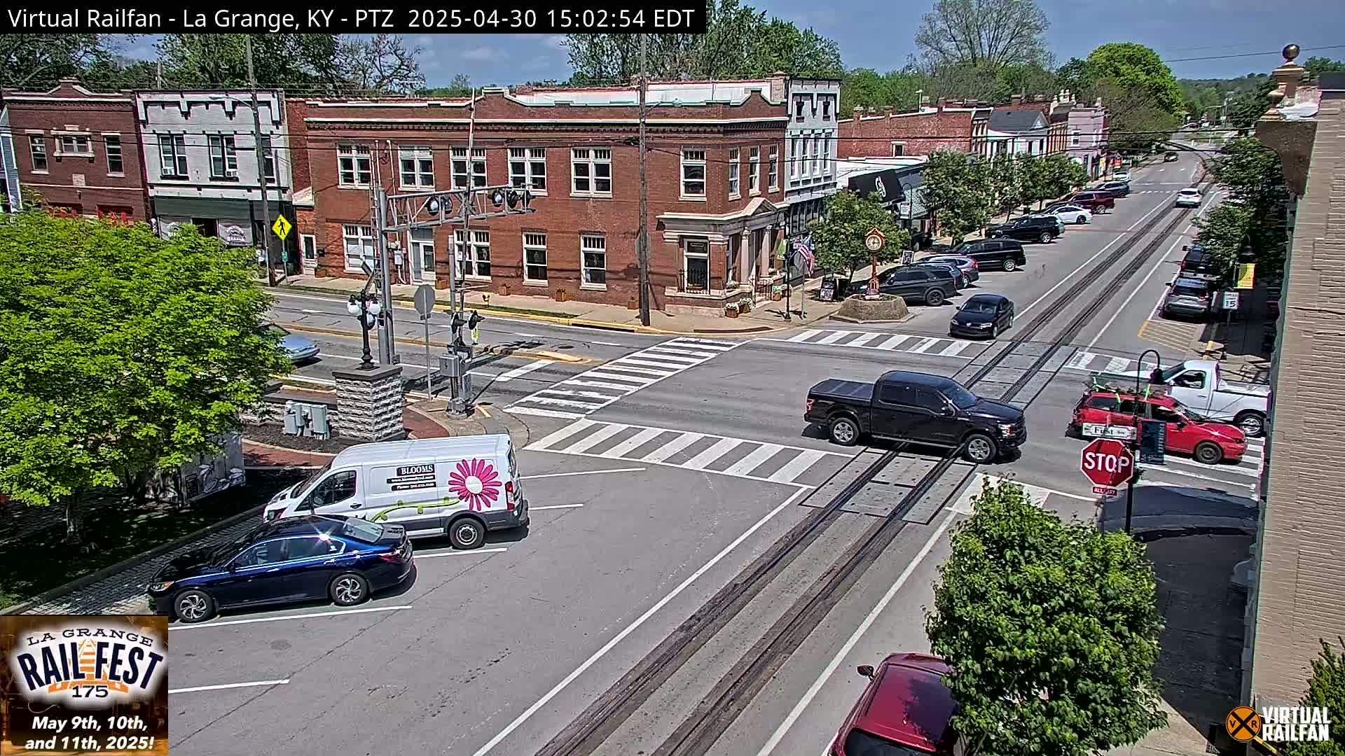 A sunny day in a small town features several cars at an intersection with train tracks running through the middle of the street, and brick buildings visible down the road.