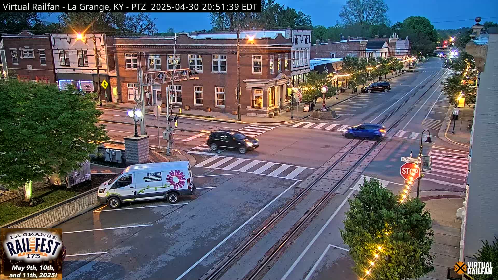 A twilight view of a town's intersection, showing several cars and a van parked near brick buildings under a mostly clear sky.