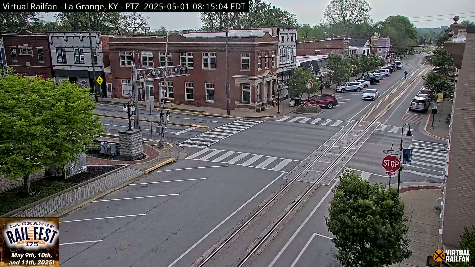 A mostly sunny view of a town's intersection with parked cars along the side of the road and a few cars driving through.