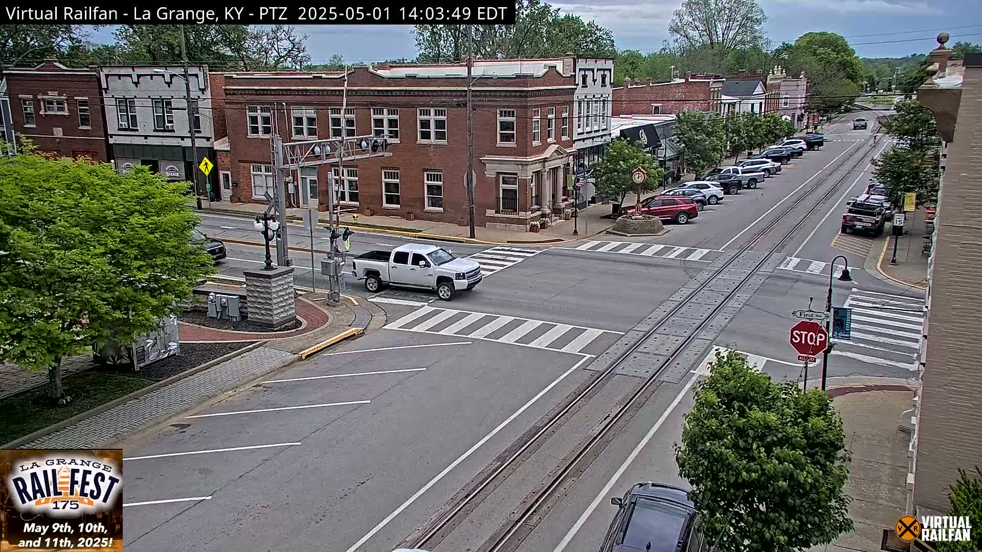 A mostly sunny day shows a partly cloudy sky over a town's intersection where a white pickup truck is crossing railroad tracks, with parked cars lining the street and brick buildings in the background.
