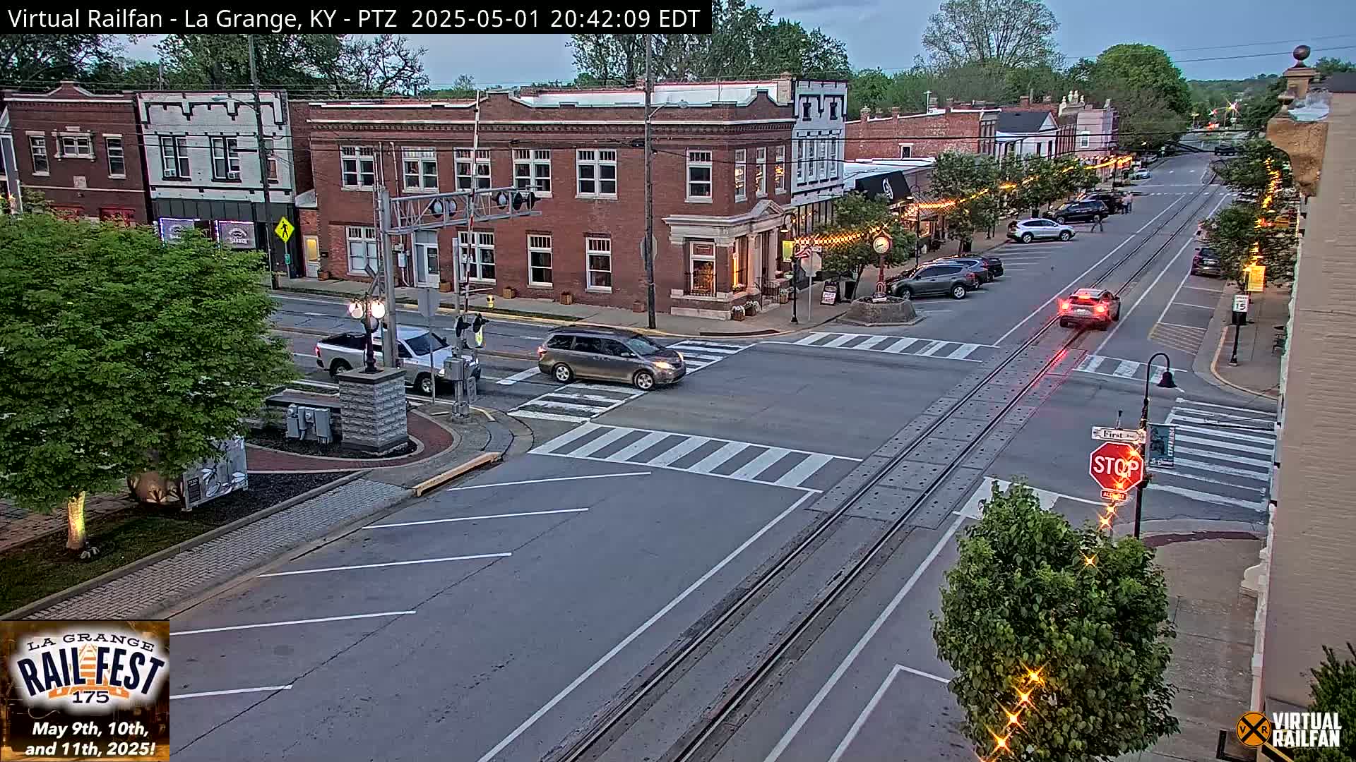 A twilight view of a town's street intersection with railroad tracks, several cars, and brick buildings.