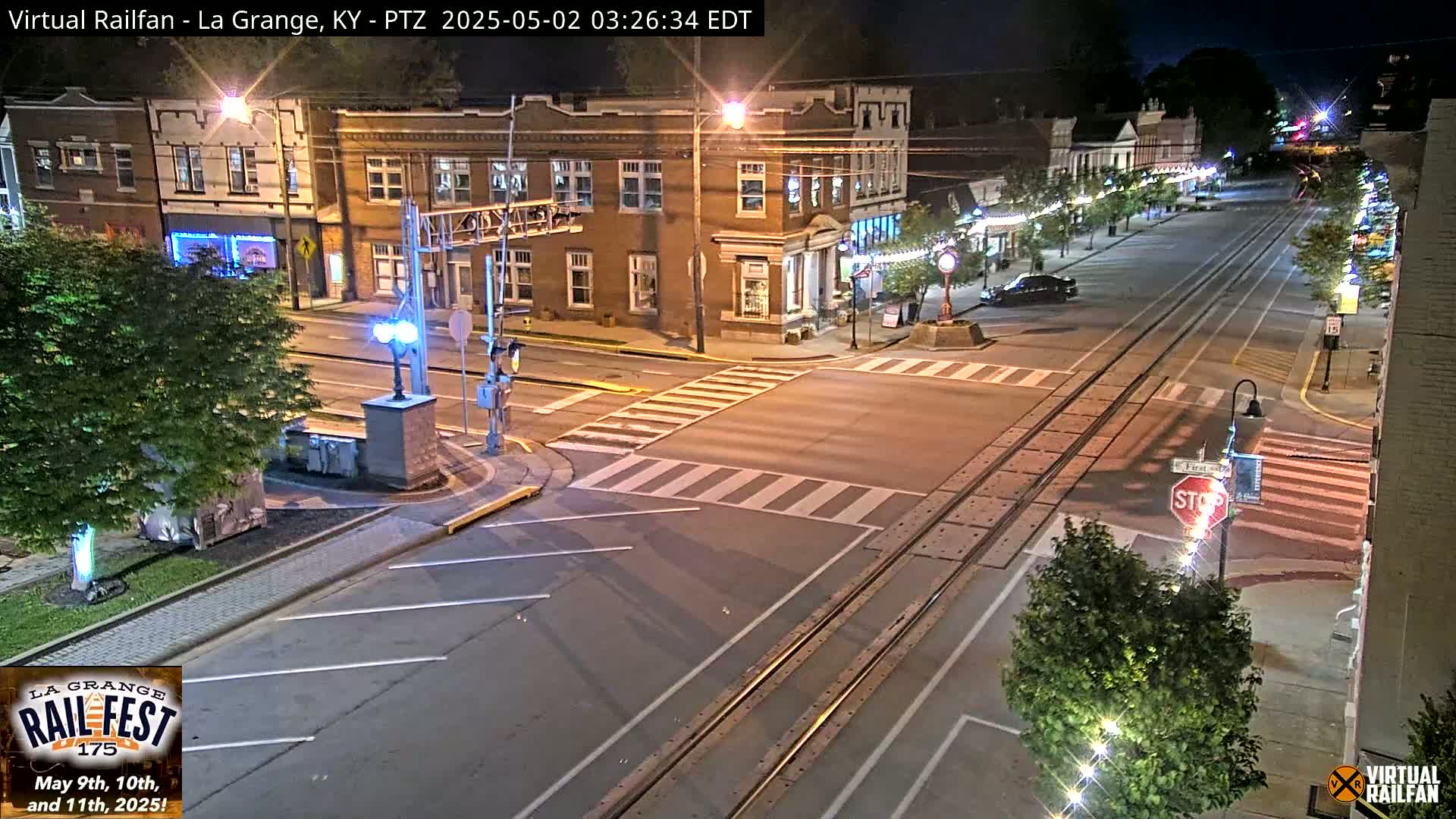 A nighttime view of a street intersection in a town with brick buildings, streetlights, and train tracks running through the middle of the road.