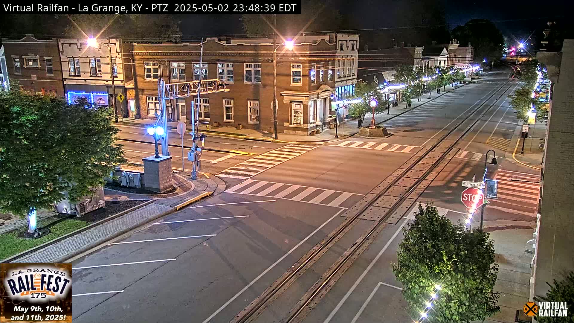 A nighttime view of a mostly empty street in a small town, with train tracks running down the middle, passing through a crosswalk at an intersection in front of brick buildings.