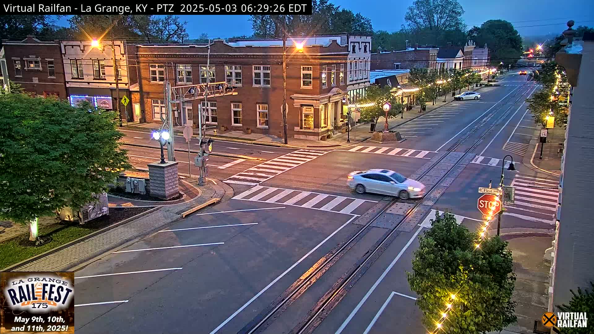 A twilight view of a town's street intersection with a car crossing railroad tracks, surrounded by brick buildings and lush green trees.