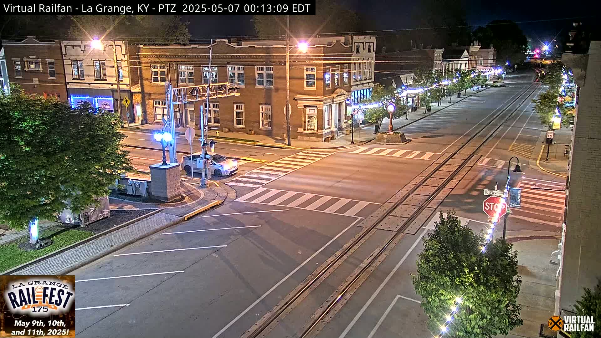A nighttime street scene shows a mostly empty road with train tracks running down the center, bordered by brick buildings and streetlights.